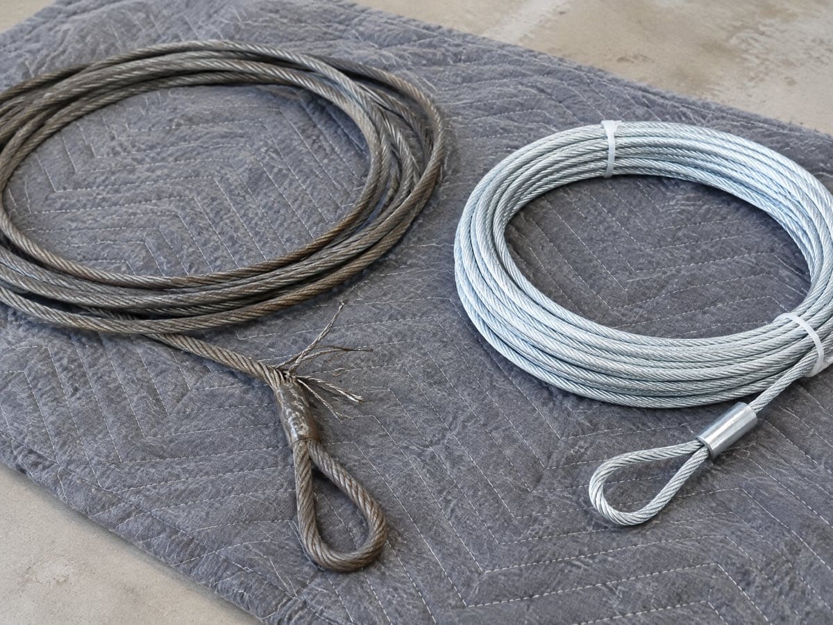 Garage door cable repair: a frayed lift cable next to a coil of new galvanized cable with a pressed loop end, on a moving blanket.