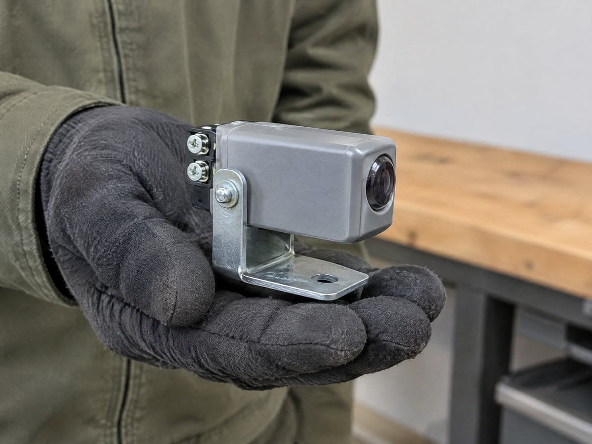Garage door sensor repair: a gloved hand holding a new photo-eye sensor in its bracket.